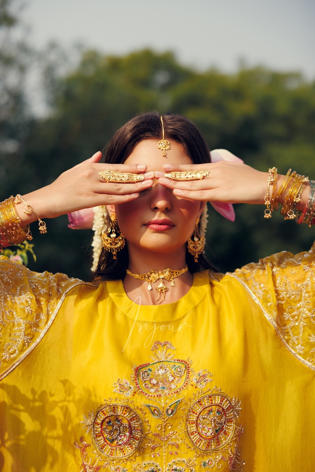 A woman outdoors wearing a bright yellow embroidered traditional outfit with ornate gold jewelry, including a headpiece, earrings, and bangles, holding her hands over her eyes.