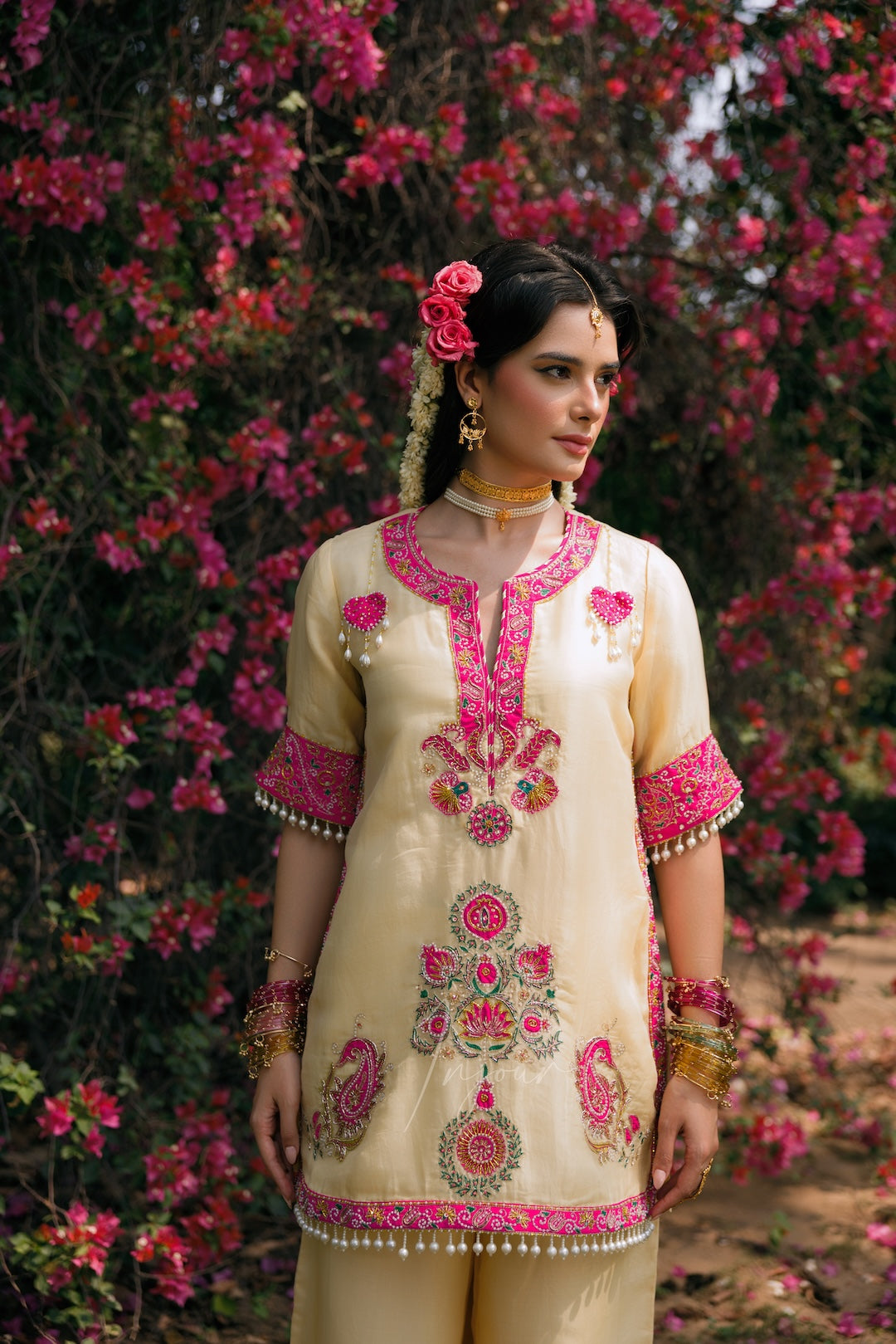 A woman wearing a cream-colored traditional kurta with vibrant pink and gold floral embroidery, standing outdoors in front of a dense background of blooming pink flowers.
