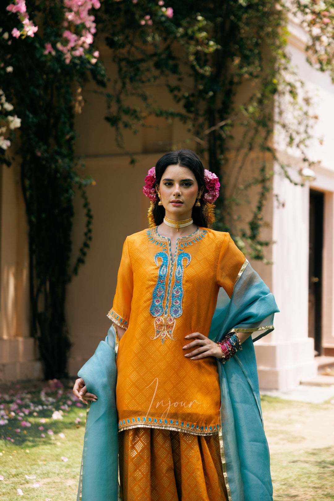 A woman stands outdoors wearing a traditional South Asian outfit with a mustard yellow kurta featuring blue and gold embroidery, a matching yellow skirt, and a teal dupatta draped over her arm. Pink flowers are in her hair, and a vine-covered building is in the background.