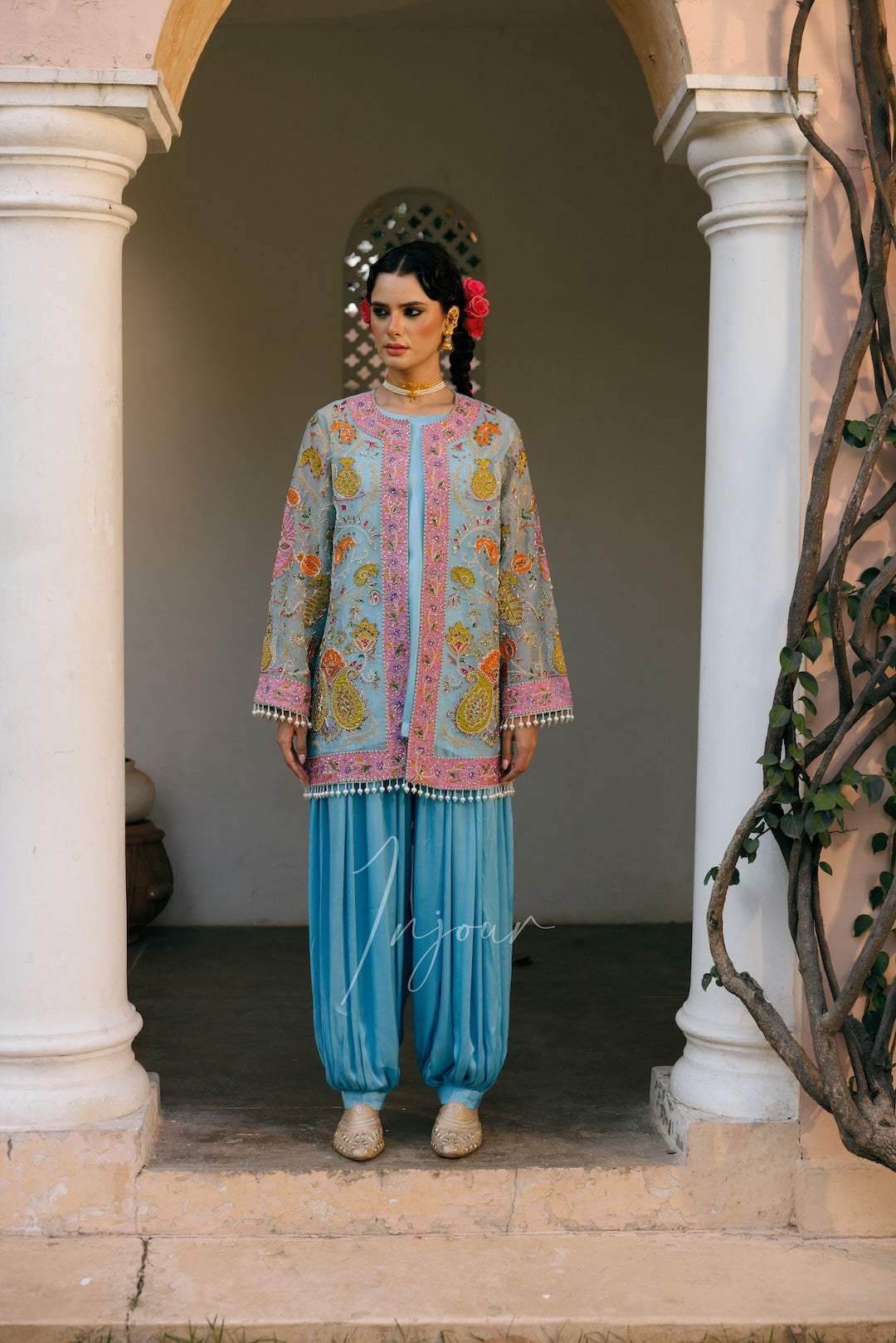 A woman stands in a stone archway doorway wearing a light blue embroidered kurta with pink and yellow paisley patterns, paired with light blue flared pants and beige shoes.