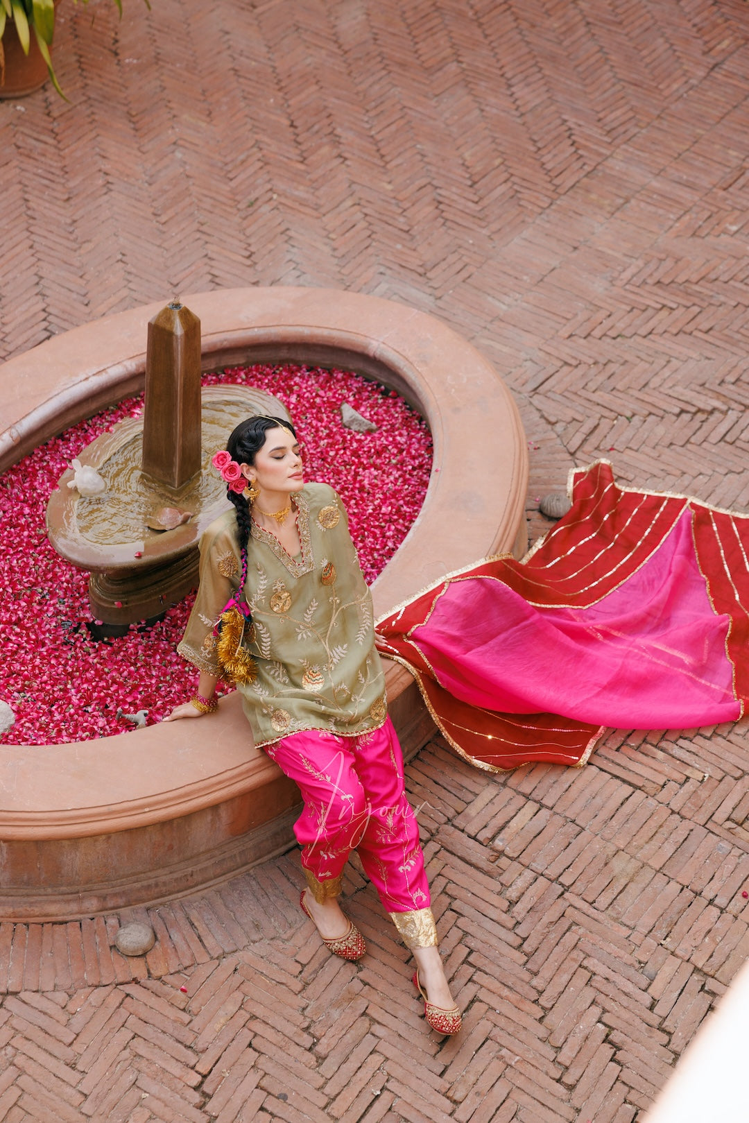 A woman wearing a traditional Indian outfit with a gold embroidered kurta, bright pink pants, and a pink dupatta sits on the edge of a circular fountain filled with red flower petals on a brick-paved courtyard.