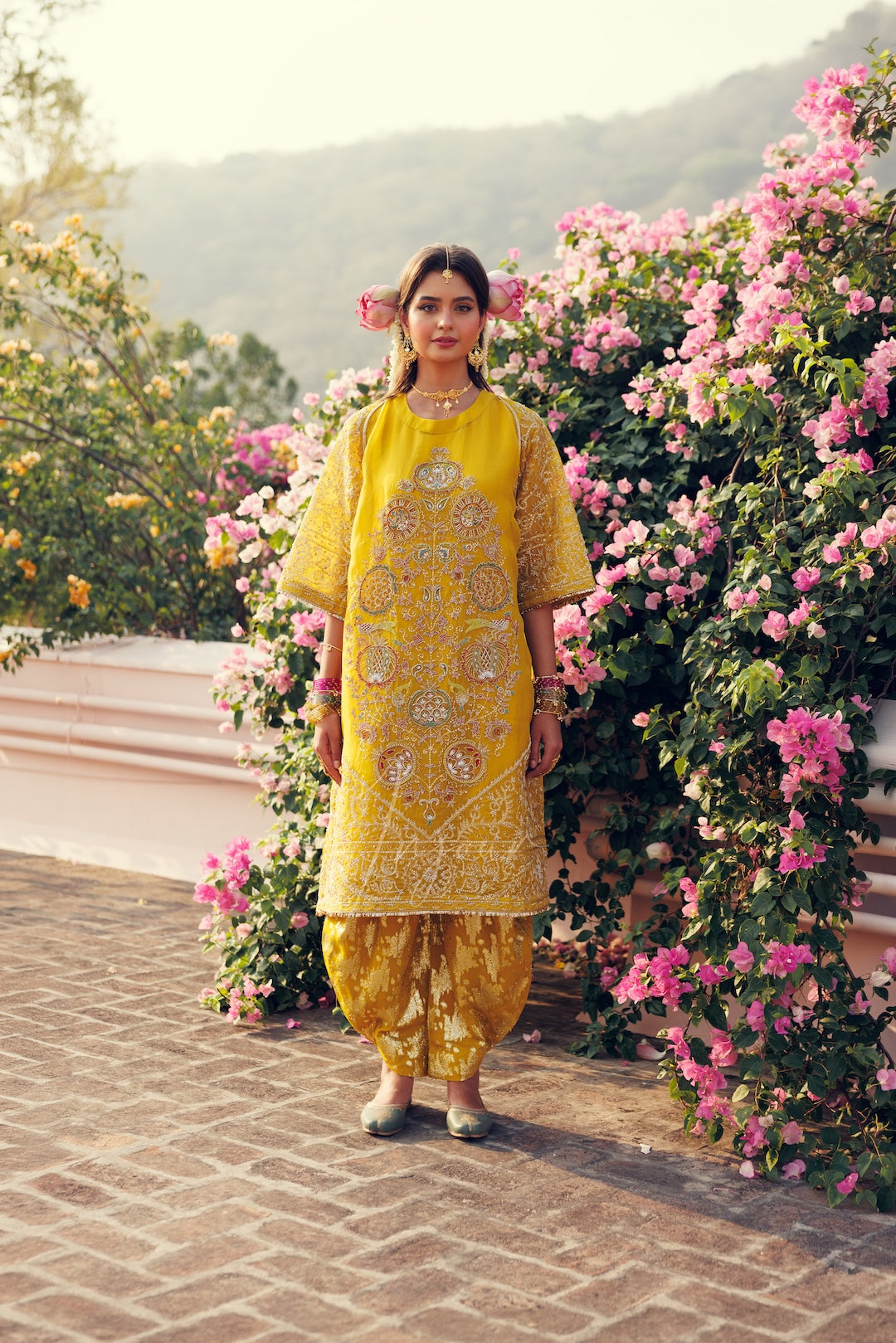 A woman stands outdoors wearing a bright yellow embroidered traditional outfit with matching patterned pants, posed in front of pink flowering bushes and a soft-focus mountain background.