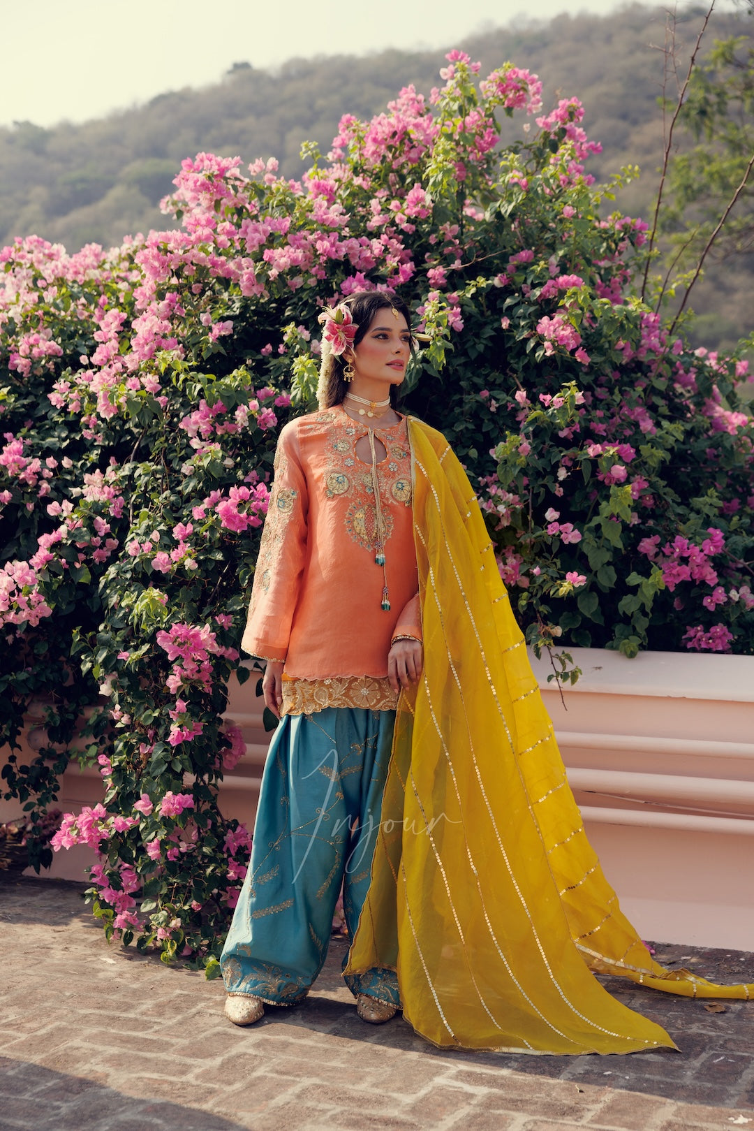 A woman stands outdoors in front of a large pink flowering bush, wearing a traditional outfit with an orange embroidered top, teal wide-leg pants, and a bright yellow sheer dupatta.