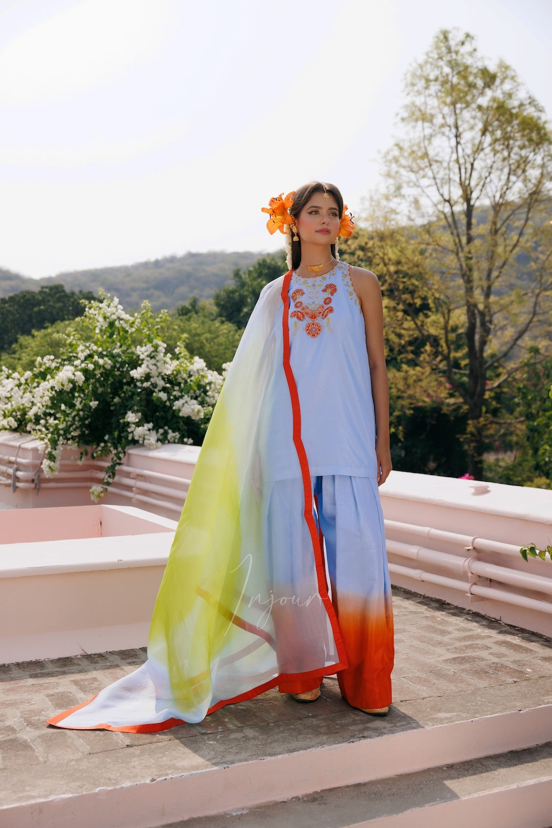 A woman stands outdoors on a terrace wearing a light blue traditional outfit with a long multicolor ombre drape in yellow, green, and orange, along with orange flower accessories in her hair.