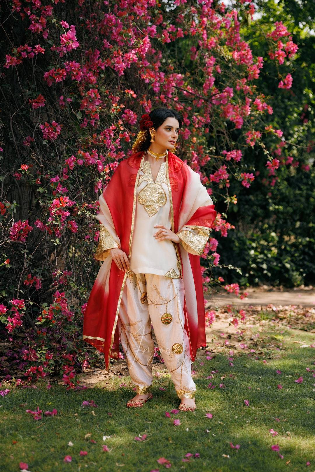 A woman stands outdoors in a garden wearing a traditional outfit with a cream kurta and pants, gold embroidery, and a red draped shawl. Pink flowers and green foliage fill the background.
