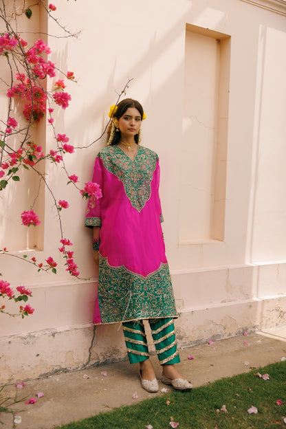 A woman models a vibrant magenta pink and green embroidered traditional kurta set with striped trousers, standing against a cream-colored stucco wall with pink flowers on the left.