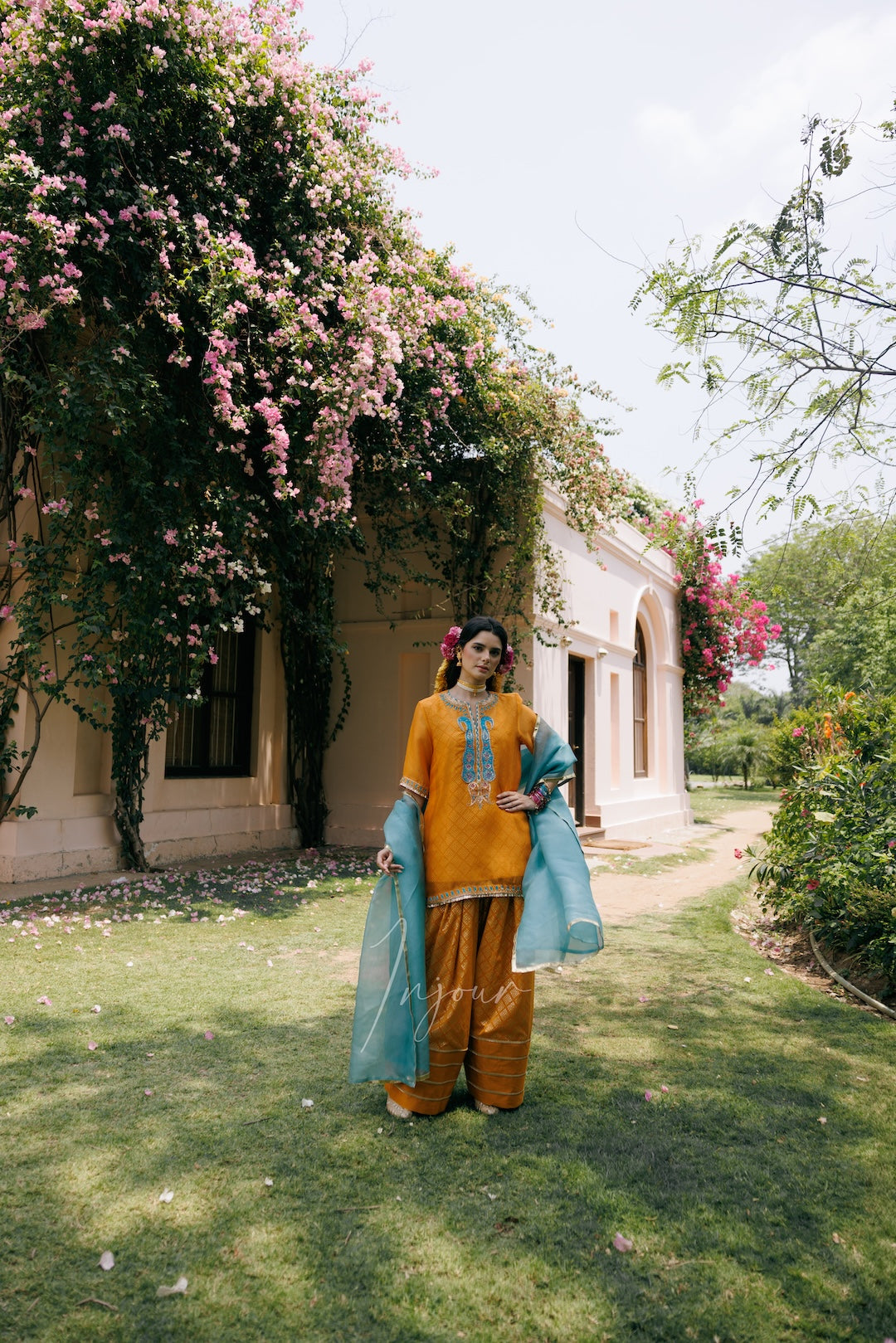 A woman wearing a traditional Indian outfit stands in a garden courtyard. The outfit consists of a mustard yellow kurta with blue embroidery, matching yellow trousers, and a light blue dupatta.