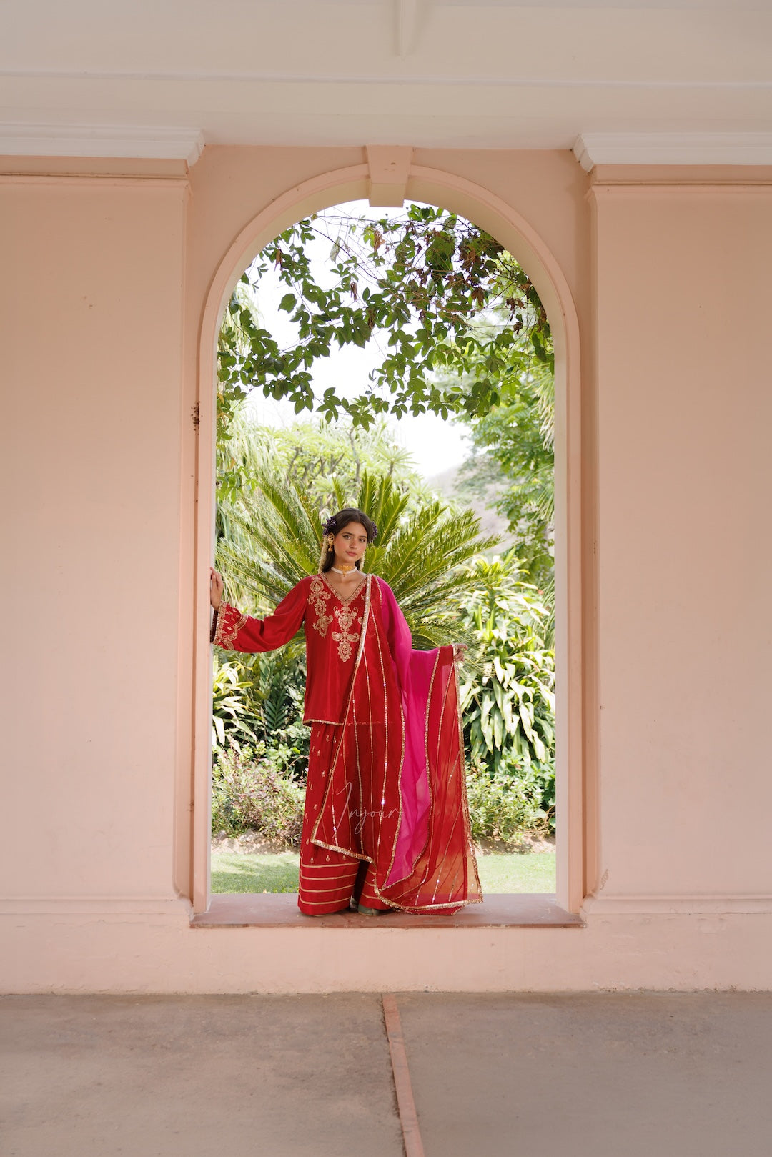 A woman wearing a traditional red and pink embroidered sari stands in the center of a peach-colored arched doorway, with lush green garden foliage visible behind her.