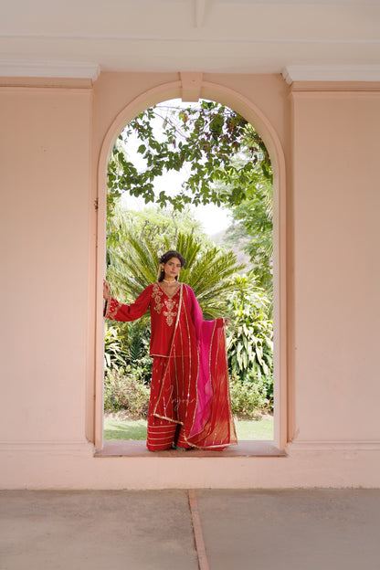 A woman wearing a traditional red and pink embroidered sari stands in the center of a peach-colored arched doorway, with lush green garden foliage visible behind her.
