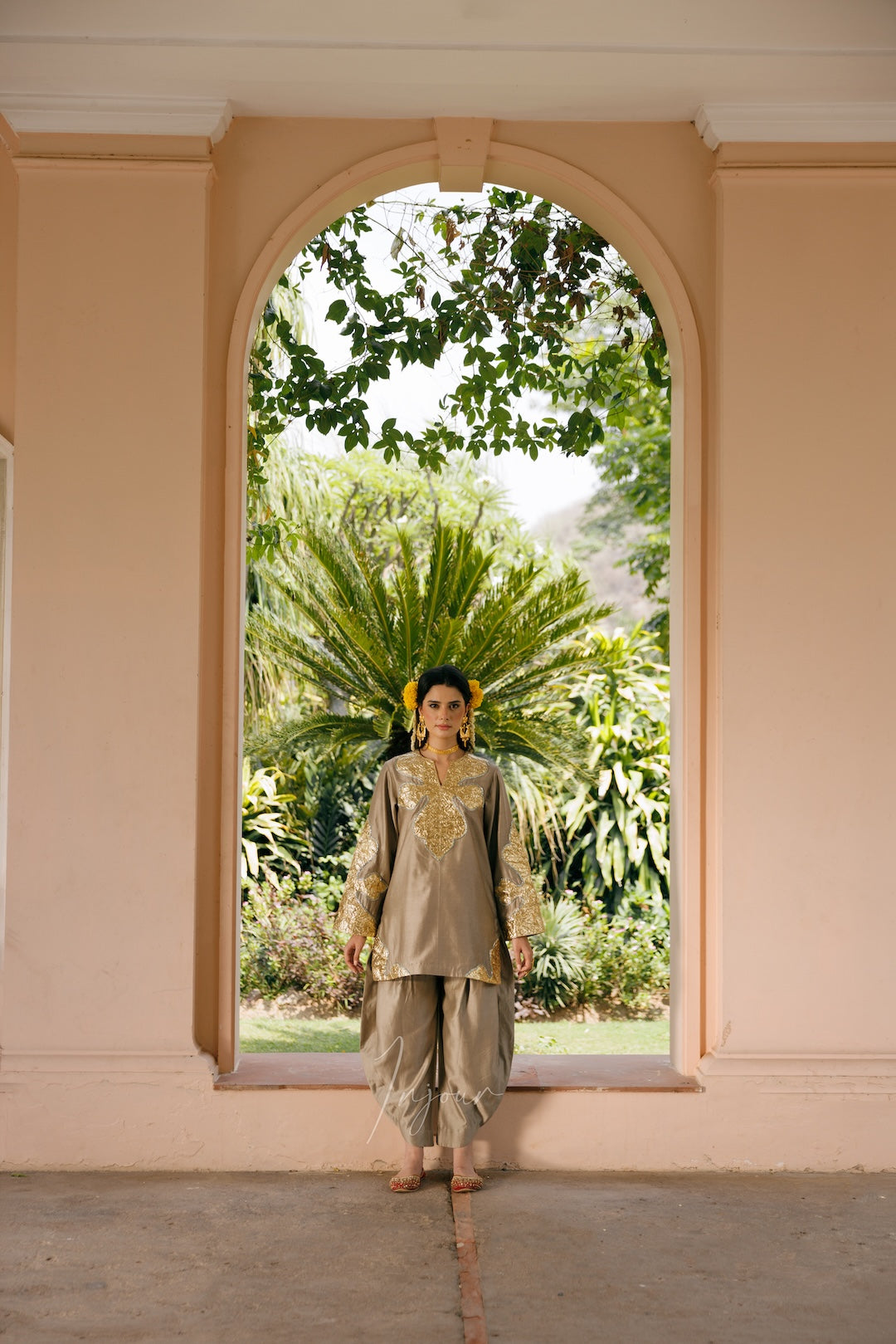 A woman stands centered in a peach-colored arched doorway, wearing a light beige traditional outfit with gold embroidery and loose draped pants. Lush green tropical foliage is visible outside the archway.