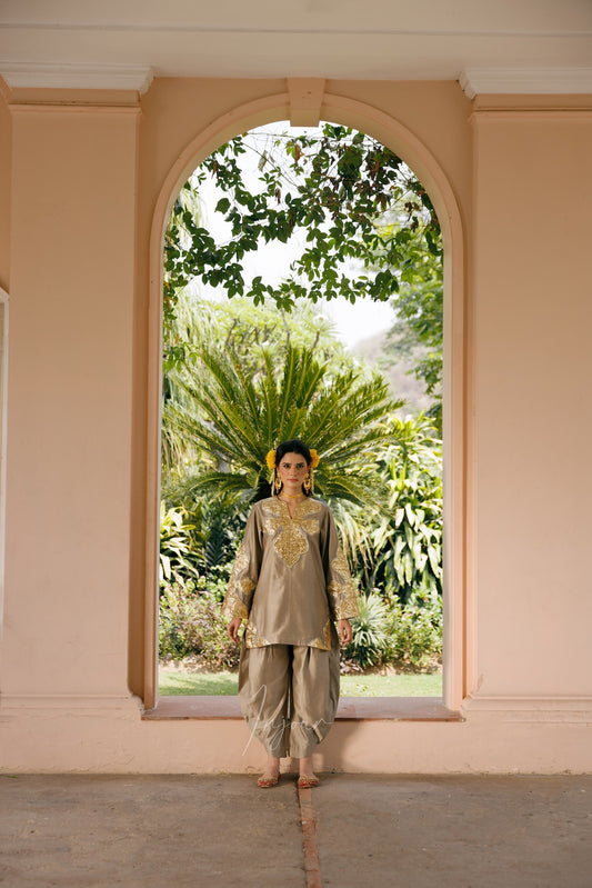A woman stands centered in a peach-colored arched doorway, wearing a light beige traditional outfit with gold embroidery and loose draped pants. Lush green tropical foliage is visible outside the archway.