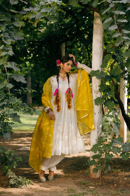 A woman stands outdoors in a garden or orchard wearing a traditional outfit with a white tunic and pants, a bright yellow draped outer layer, and pink decorative garlands in her hair.