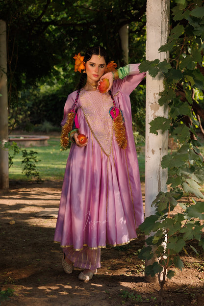 A woman stands outdoors in a garden wearing a flowing lavender-pink traditional dress with gold embellishments, floral hair ornaments, and colorful draped accents. Trees, vines, and a dirt path are visible around her.