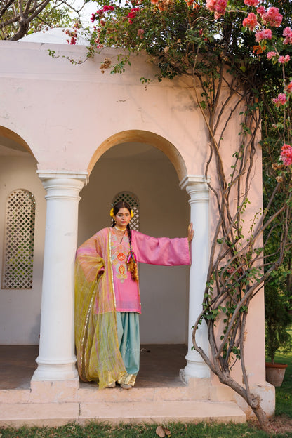 A woman wearing a traditional Indian outfit with a pink top, teal skirt, and yellow-green dupatta stands in the center of a white arched corridor decorated with climbing pink flowers.