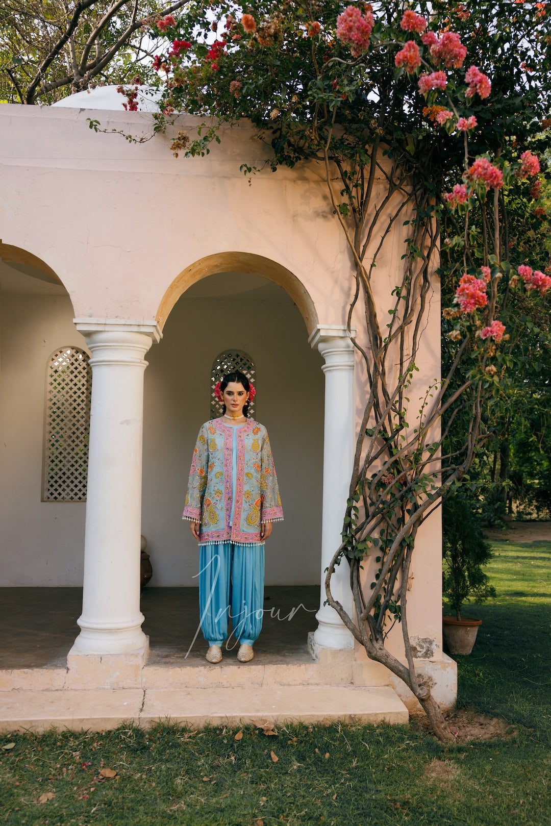 A woman stands centered under a white arched pavilion in a garden, wearing a light blue traditional outfit with colorful embroidered details and a matching headpiece. Pink flowers and climbing vines frame the scene, with green grass visible around the structure.