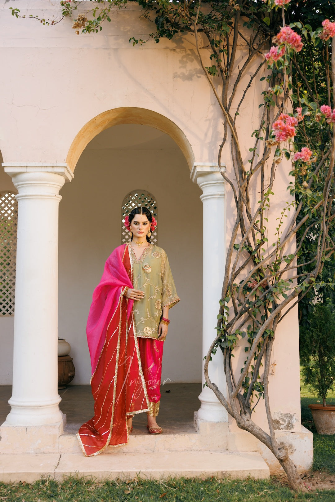 A woman stands in the center of a white arched doorway wearing a traditional outfit with a bright pink dupatta, a gold embroidered kurta, and a red skirt. Pink flowering vines climb along the right side of the structure.