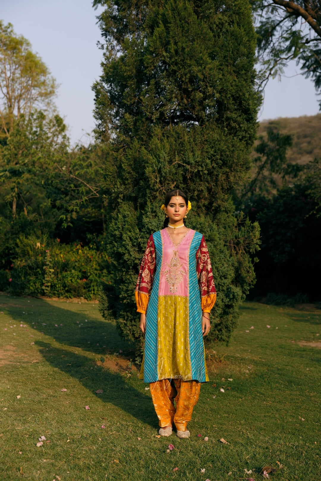 A woman stands outdoors on a grassy lawn in front of a tall dark green tree, wearing a colorful traditional outfit with a pink and yellow embroidered kurta, turquoise and yellow border, and orange-yellow pants.