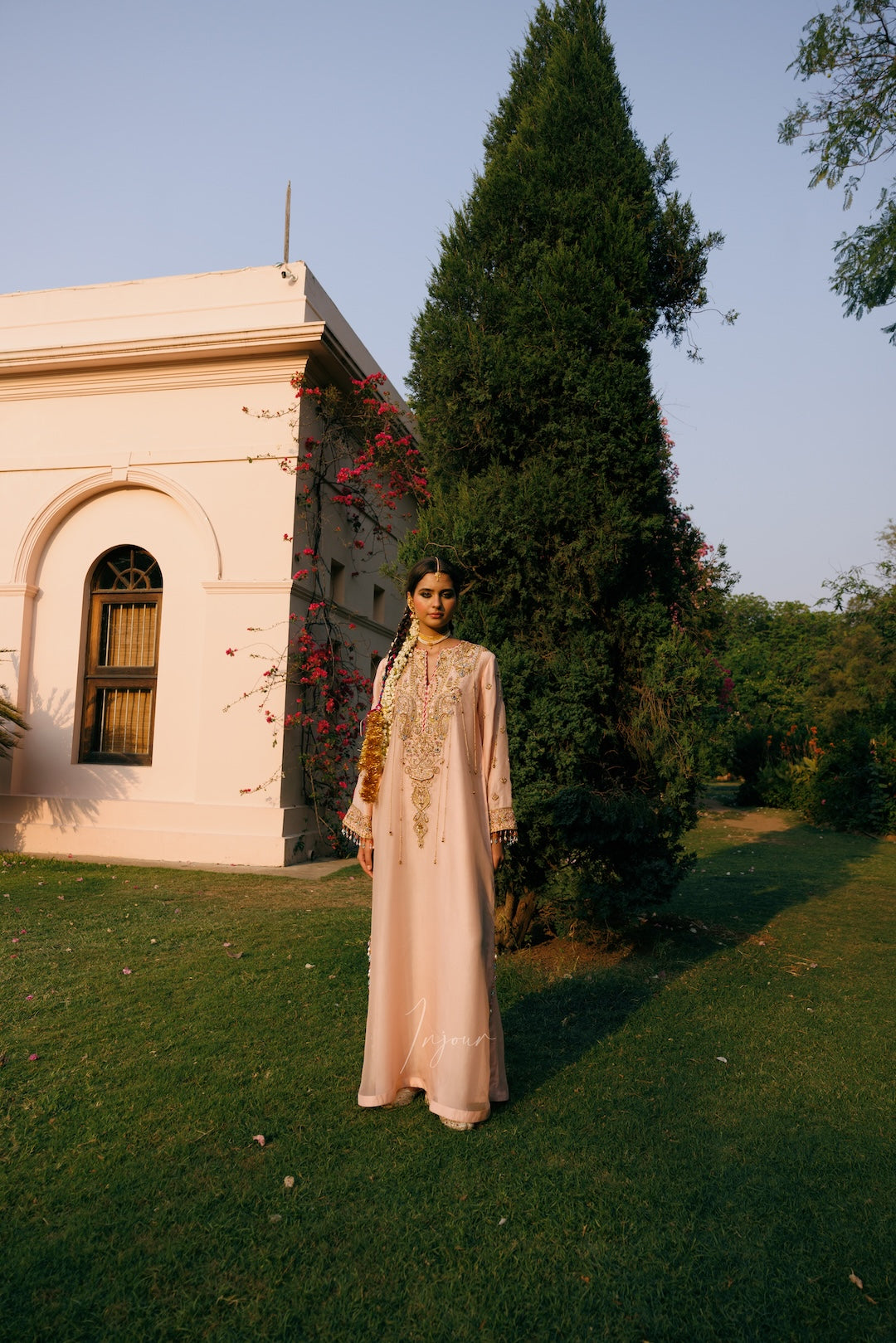A woman stands on a grassy lawn wearing a long, light pink traditional dress with gold embroidery, posed in front of a pale pink building and a tall green tree.