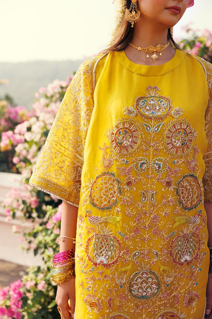A woman wearing a bright yellow embroidered kurta with intricate multicolor floral and geometric patterns, styled with gold jewelry and bangles, set against a soft-focus background of pink flowers.