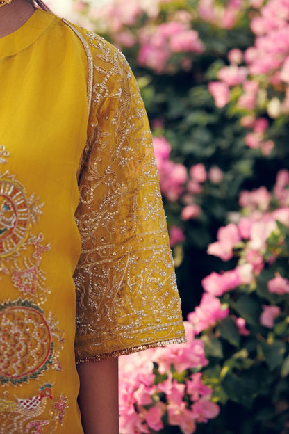 A close-up of a mustard yellow embroidered kurta with ornate silver and gold beadwork on the sleeve, worn by a model in front of a soft-focus background of pink flowers and green foliage.
