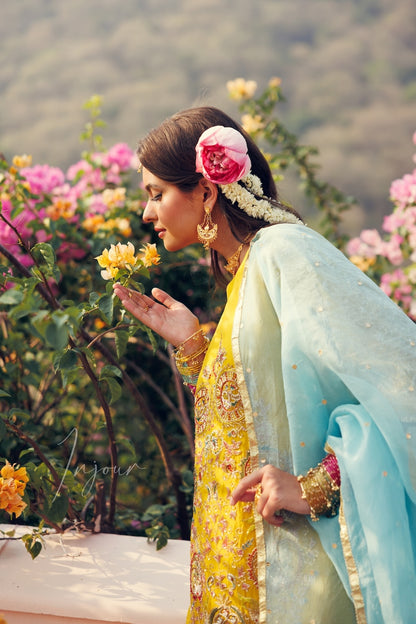 A woman in traditional South Asian attire wearing a yellow embroidered kurta and a light blue dupatta, posing outdoors among pink flowers and smelling yellow blossoms. She has a pink and white flower in her hair and is wearing gold jewelry.