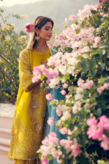 A woman wearing a bright yellow embroidered traditional outfit with a turquoise dupatta, standing outdoors beside a large bush of pink flowers.