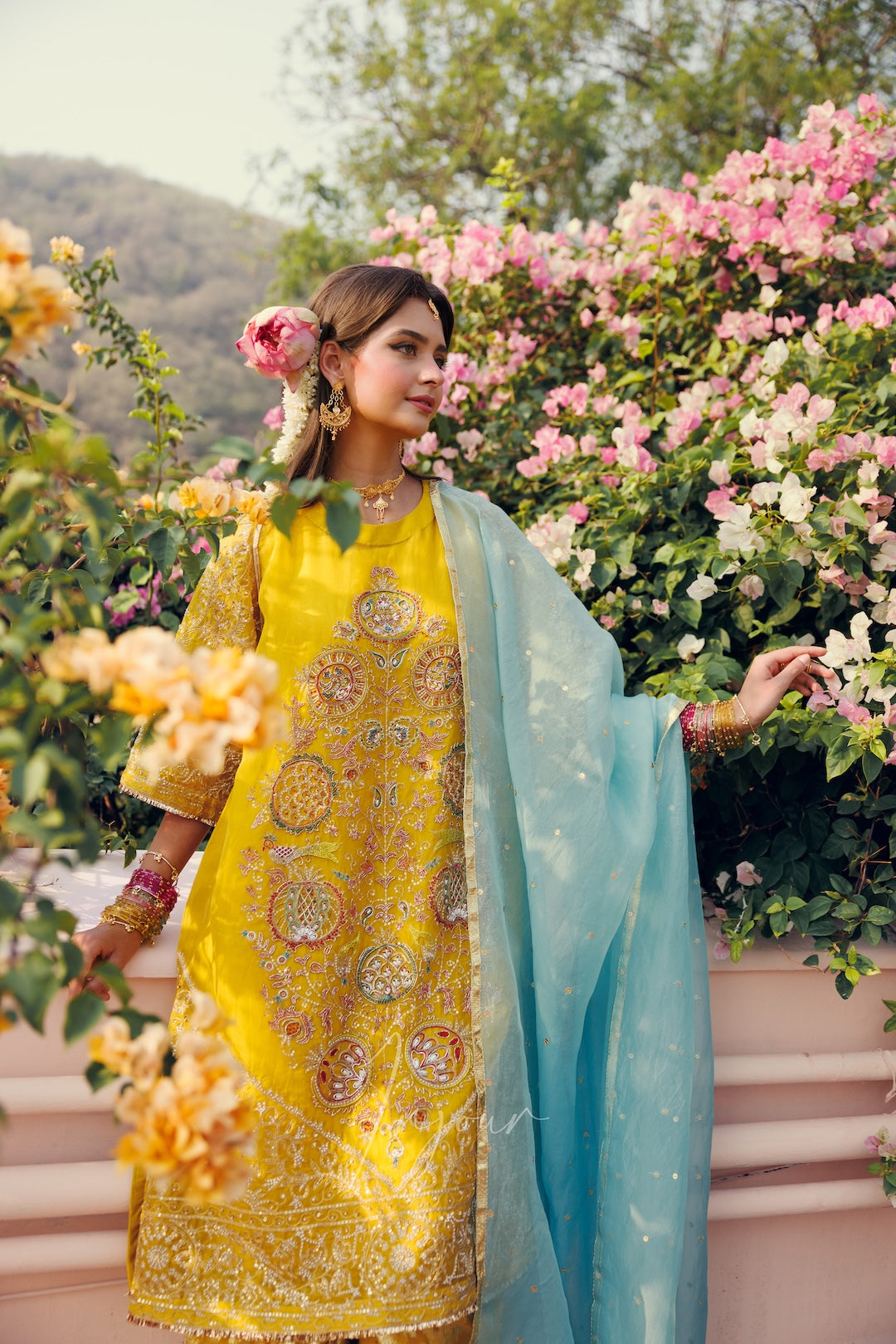 A woman wearing a traditional Indian outfit in bright yellow with intricate gold embroidery and a light blue dupatta, standing outdoors in front of pink flowers and green foliage.