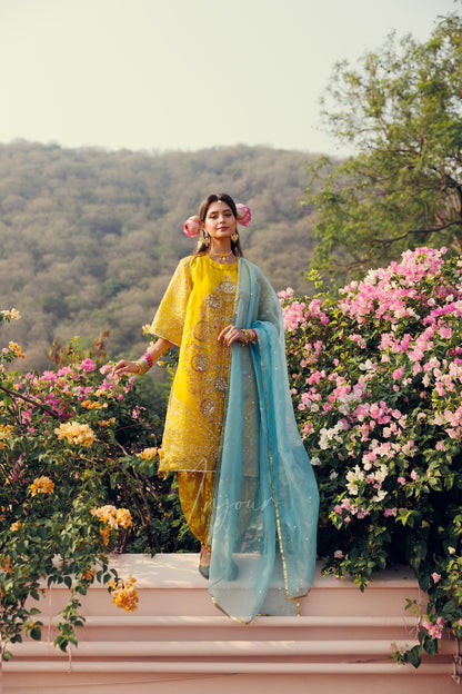 A woman wearing a traditional Indian outfit stands on a pink terrace surrounded by pink and yellow flowers, with hills and trees in the background. She is wearing a yellow kurta with gold embroidery, matching yellow trousers, and a light blue dupatta.