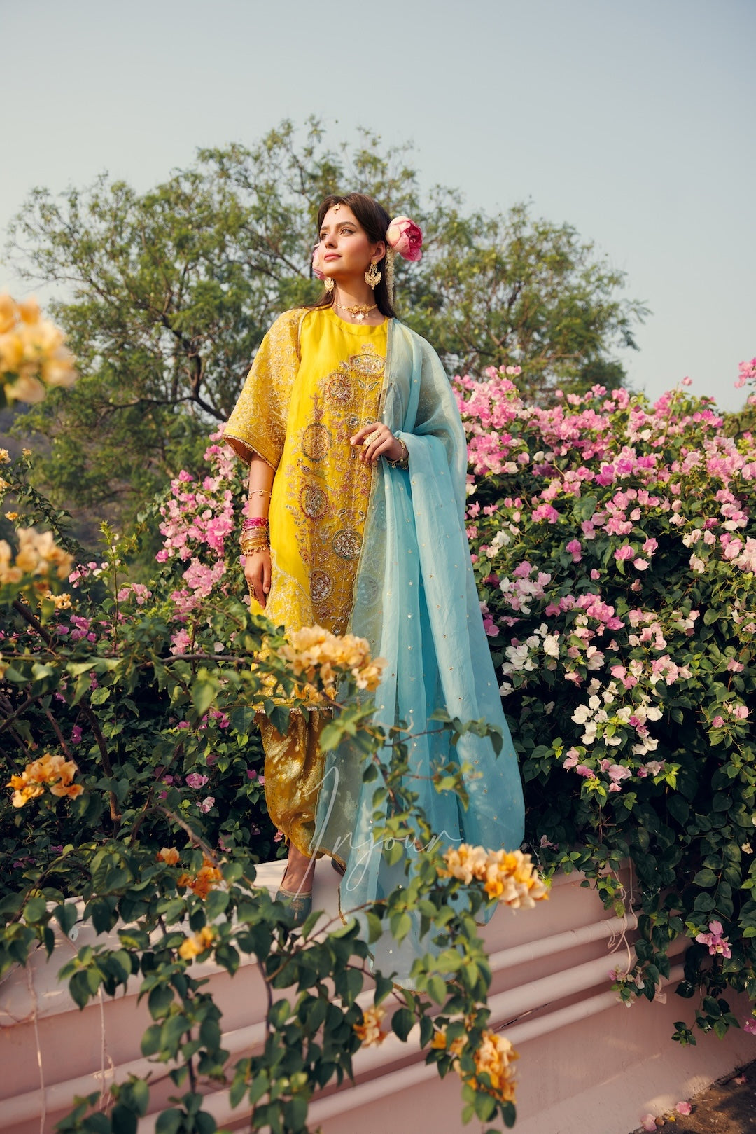 A woman stands outdoors in a lush garden wearing a traditional outfit with a yellow embroidered kurta, matching yellow trousers, and a light blue dupatta draped over her shoulder.