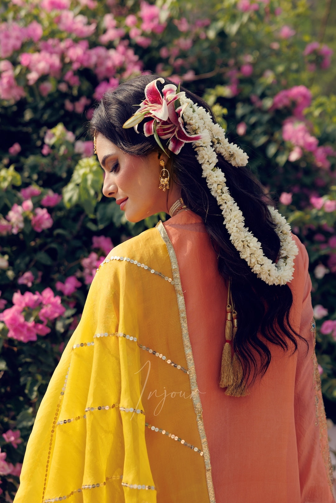 A woman shown from the back wearing a traditional outfit with a bright yellow dupatta featuring silver mirror work borders, a peach-colored kurta, and a floral garland in her hair, set against a background of blooming pink flowers.