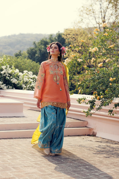 A woman wearing a traditional Indian outfit with an orange embroidered kurta, teal blue pants, and a yellow dupatta, standing outdoors on stone steps with flowering trees in the background.