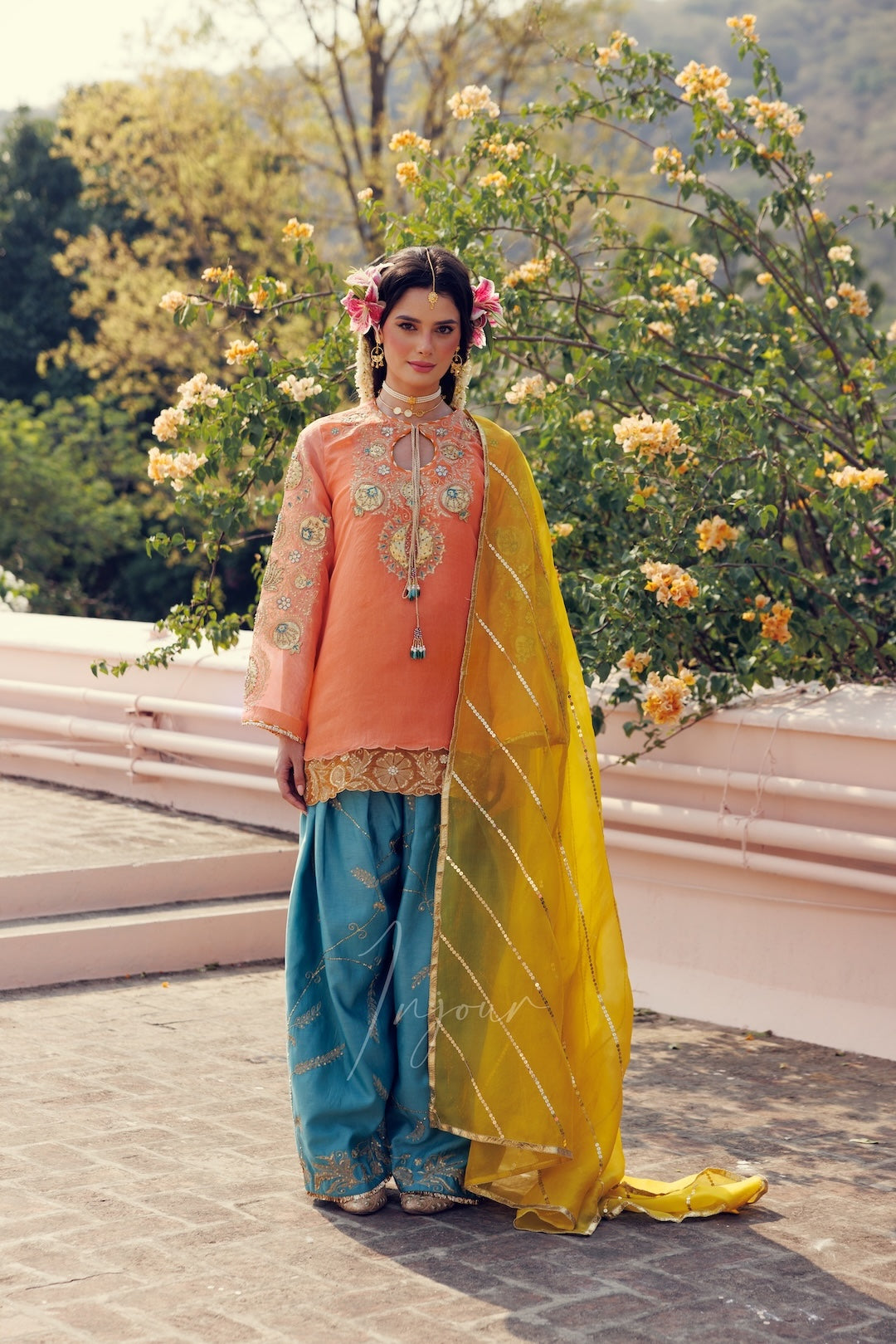 A woman stands outdoors wearing a traditional outfit with a peach embroidered kurta, teal wide-leg pants, and a bright yellow dupatta with silver stripes. She is posed in front of flowering bushes and a stone wall.