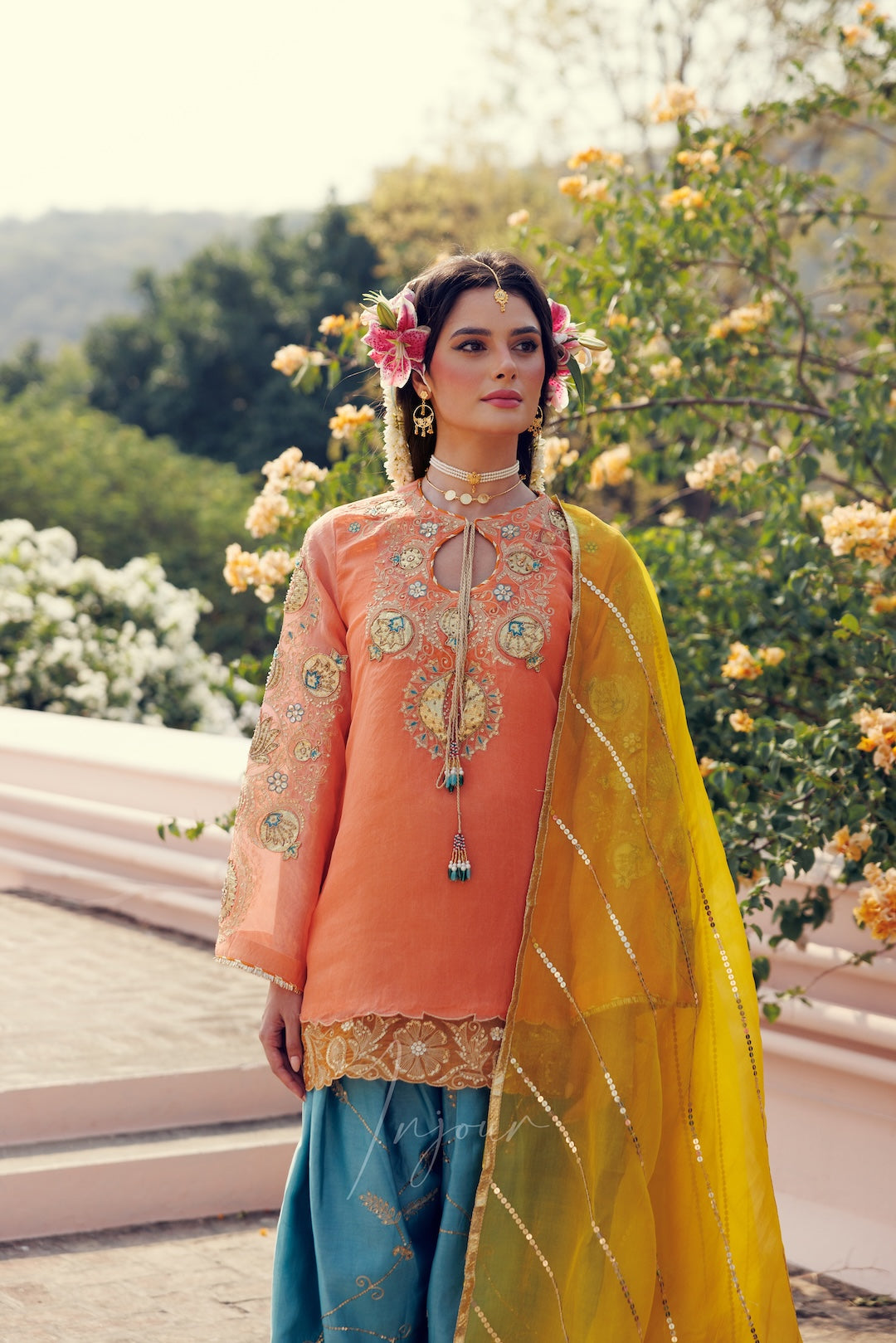 A woman models a traditional three-piece salwar kameez suit. The outfit features a peach-orange embroidered kameez top with a matching tie, teal blue salwar pants, and a bright yellow dupatta with gold zari detailing.
