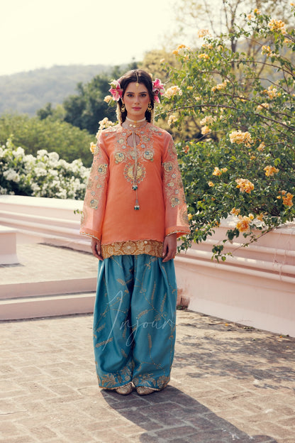 A woman stands outdoors wearing a traditional outfit with a peach embroidered top and teal wide-leg pants with gold detailing. The background features a stone terrace, greenery, and flowering bushes.