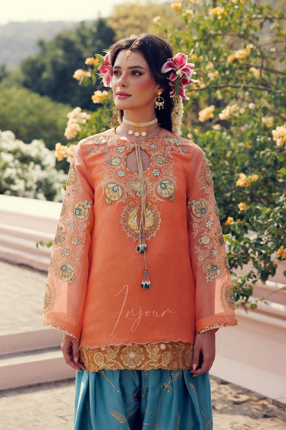 A woman wearing a peach-orange embroidered traditional kurta with sheer sleeves and a gold border, paired with a blue skirt, standing outdoors in front of flowering bushes.