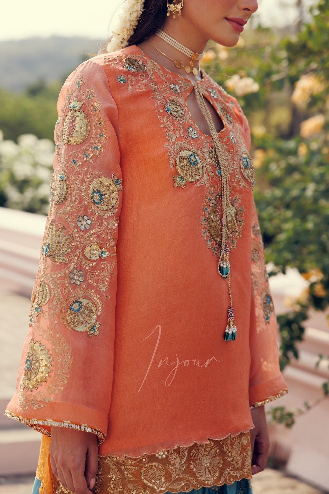 A woman wearing a peach-orange embroidered traditional kurta with ornate gold and multicolor embellishments, shown in an outdoor garden setting.