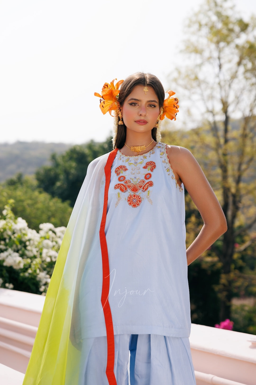 A woman wearing a light blue traditional outfit with orange floral embroidery on the chest, a bright yellow-green dupatta with a red stripe, and orange flower accessories in her hair, set against a blurred outdoor garden background.
