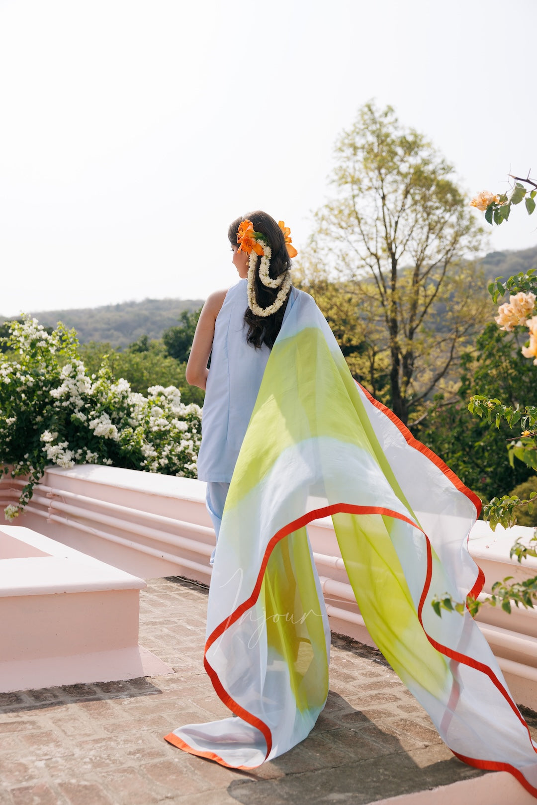 A woman stands outdoors on a terrace wearing a flowing sheer white sari with bright yellow-green panels and red-orange border trim. The background features green trees, hills, and pink architectural structures under a bright sky.