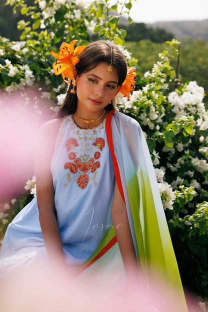A woman wearing a light blue embroidered traditional outfit with a multicolor draped scarf, seated outdoors among green foliage and white flowers. She has orange flowers in her hair and a gold headpiece.
