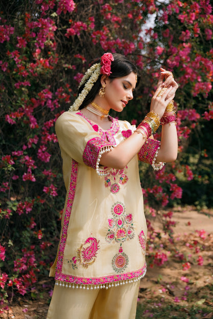 A woman wearing a cream-colored traditional South Asian outfit with vibrant pink and green floral embroidery, standing outdoors in front of a dense background of blooming pink flowers.