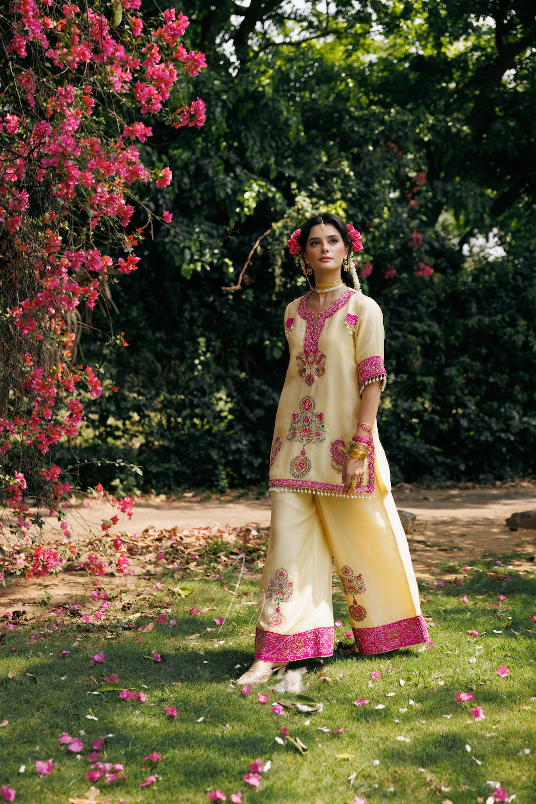 A woman stands outdoors in a garden wearing a cream-colored traditional outfit with pink and gold embroidery. The scene includes green grass, trees, and a large bush of pink flowers on the left.