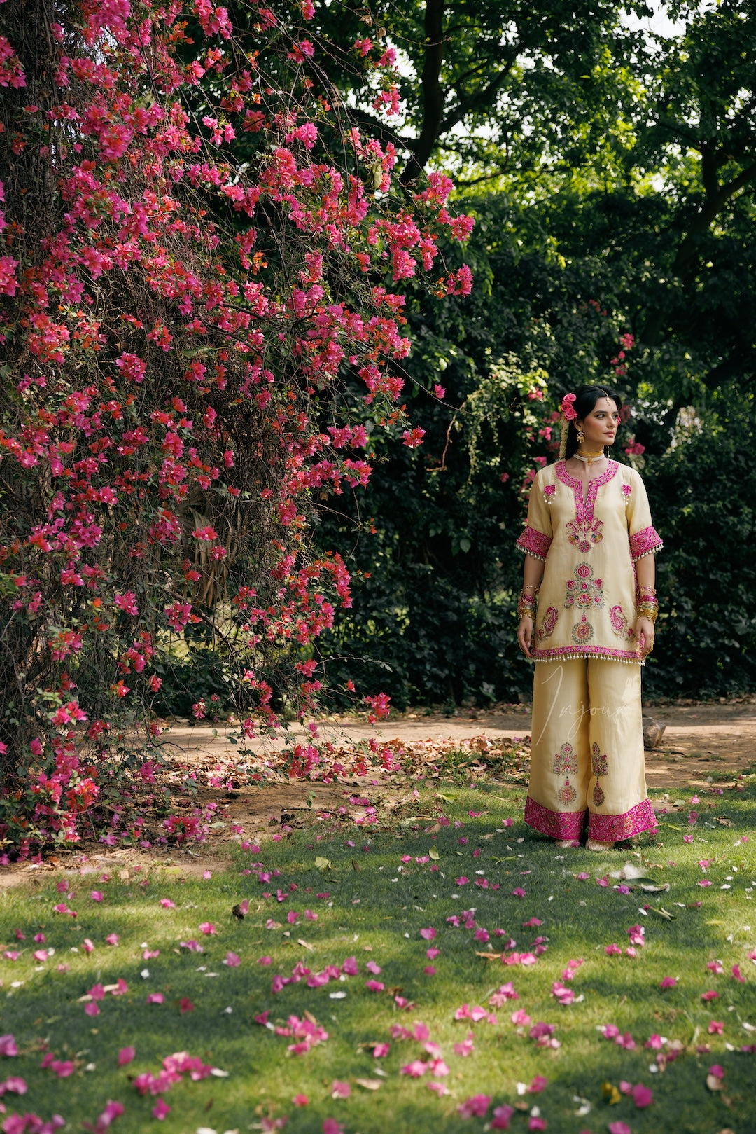 A woman stands outdoors on a grassy lawn under a large flowering bush with pink blossoms. She is wearing a light beige traditional outfit with pink embroidery and matching pink-trimmed pants.