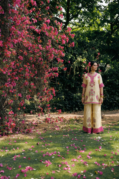 A woman stands outdoors on a grassy lawn under a large flowering bush with pink blossoms. She is wearing a light beige traditional outfit with pink embroidery and matching pink-trimmed pants.