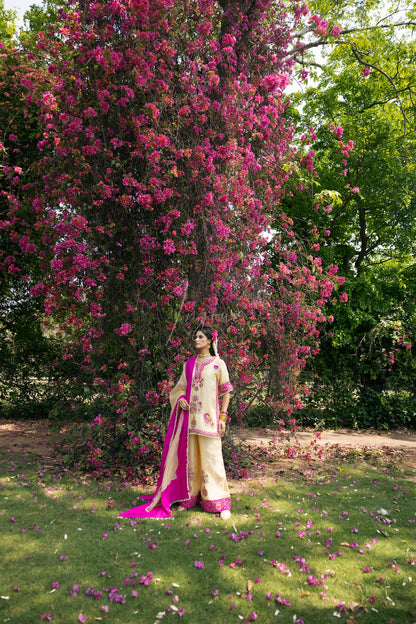 A woman stands outdoors under a large flowering tree with pink blossoms, wearing a traditional outfit with a cream-colored top and skirt, a bright pink dupatta, and pink shoes. Green grass and fallen petals are visible on the ground.