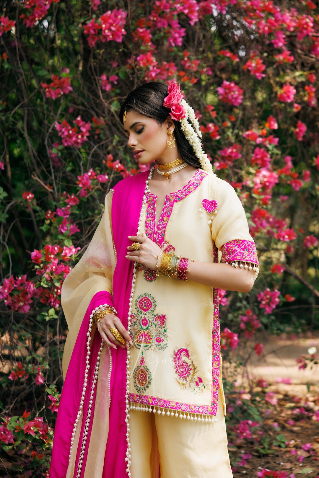 A woman wearing a traditional Indian salwar kameez outfit in cream and bright pink, standing outdoors in front of a background of blooming pink flowers. The outfit features intricate embroidery on the kameez front and sleeves, and a matching pink dupatta.