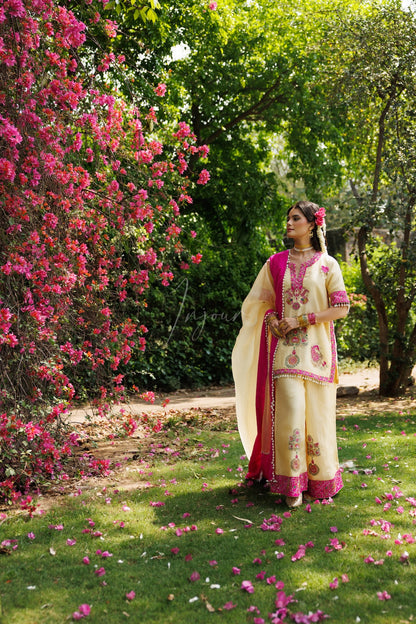 A woman wearing a cream and pink embroidered traditional outfit stands on a grassy garden path with pink flowers and green trees in the background.