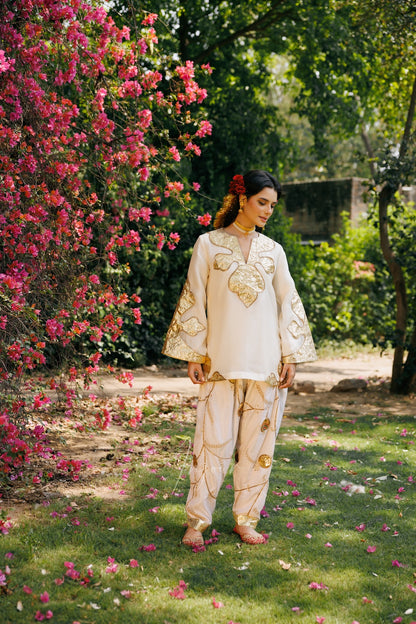 A woman stands outdoors in a garden wearing a cream-colored traditional outfit with gold embroidery. The setting features green grass, trees, and a large bush of pink flowers on the left.