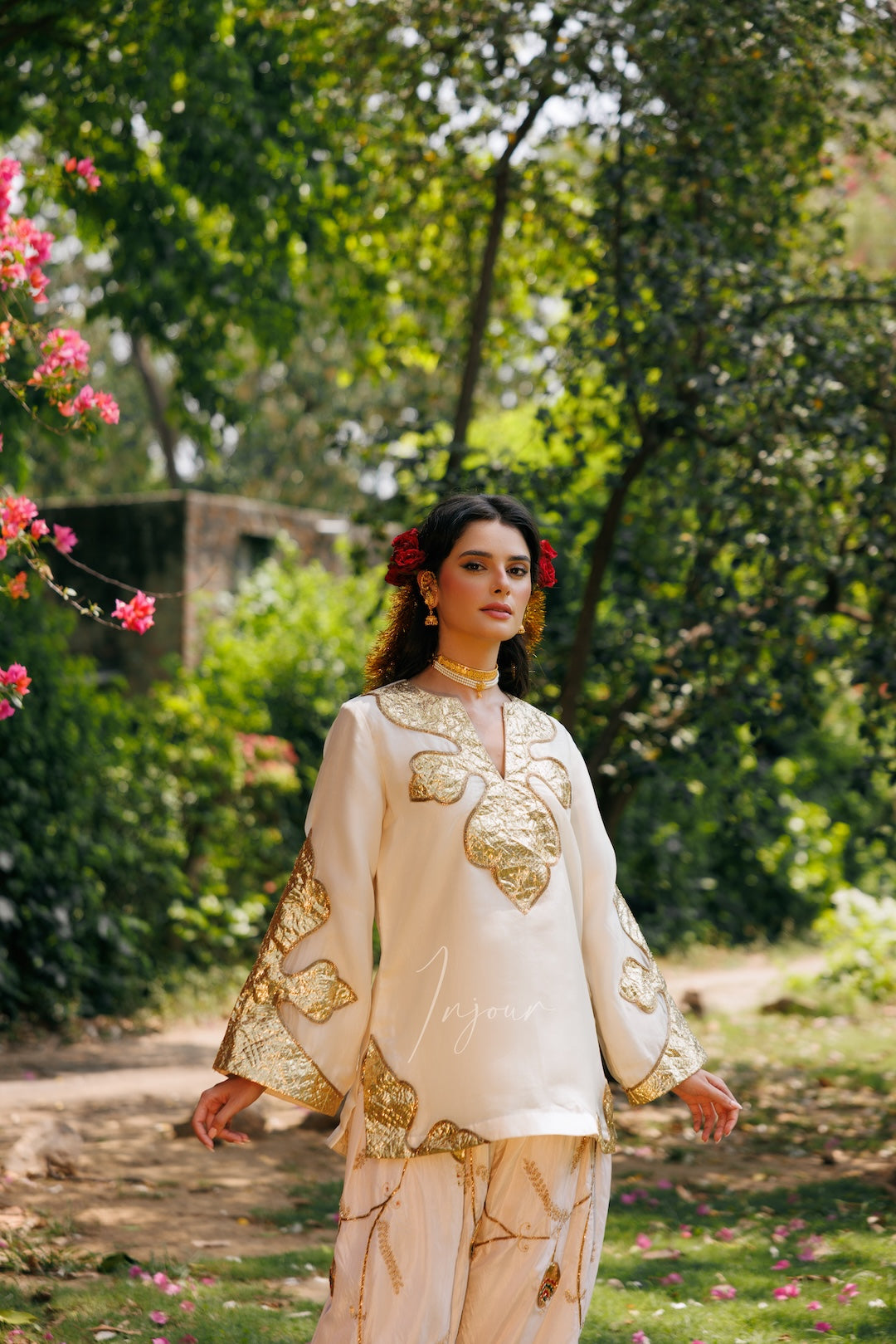 A woman stands outdoors in a garden wearing a cream-colored traditional outfit with gold embroidery and matching pants. The background features lush green foliage and trees.