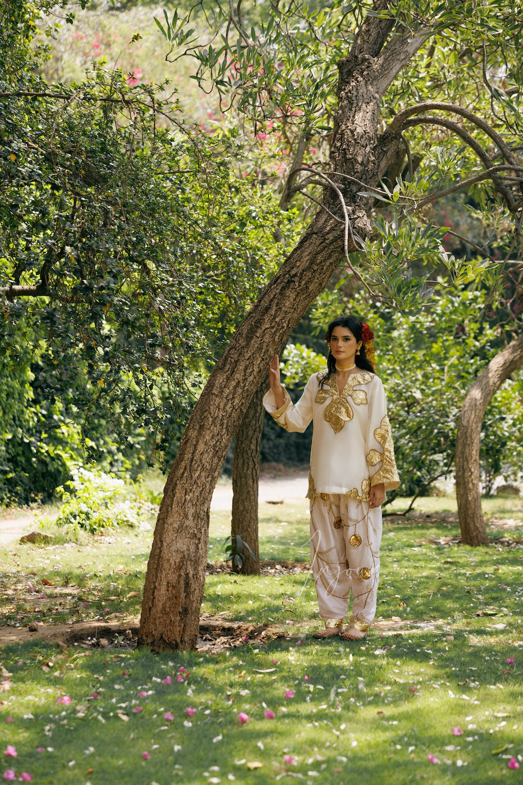 A woman wearing a cream and gold embroidered traditional outfit stands under a tree in a sunlit garden.