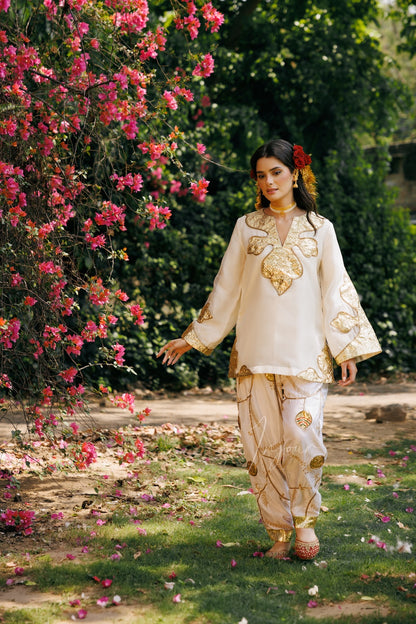A woman wearing a cream-colored traditional outfit with gold embroidery walks along a garden path next to a lush bush of pink flowers.